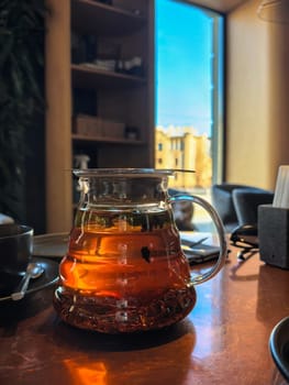 Saint-Petersburg, Russia - 04.16.2024. Glass teapot filled with brewed tea sits on a table, capturing sunlight next to a window in a cafe. Photo taken with a smartphone.