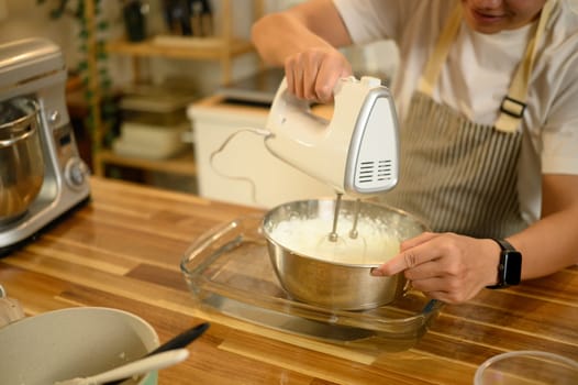 Man mixing cream with an electric beater in stainless steel bowl during a home baking session.
