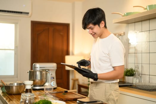Asian man in kitchen checking a recipe on digital tablet while preparing whipped cream.