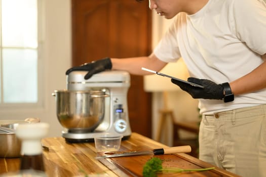 Asian man in kitchen checking a recipe on digital tablet while preparing whipped cream.