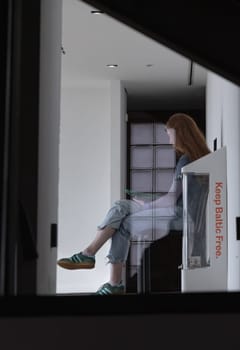 Newcastle, UK - Apr 12, 2025 - Looking through the glass at a long-haired young woman wearing T-shirt, jeans, sneakers sitting reading a book on bench in Baltic Flour Mills is Centre for Contemporary Art at Gateshead Quayside by the River Tyne. use it as your Wallpaper, Poster and Space for text, Selective focus.