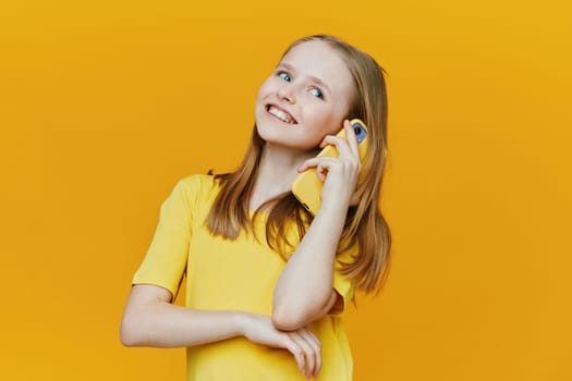 A cheerful young girl in a bright yellow shirt enjoys speaking on a yellow phone, representing happiness, youthful connection, and modern technology against a vivid yellow backdrop