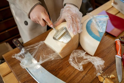 Close-up of the hands of a vendor cutting cheese at a farmers market. The vendor uses a slicer with a wooden handle to cut a thin slice of cheese surrounded by cheese wheels for a customer.