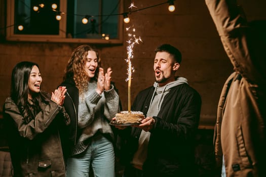 A group of friends joyfully celebrates a birthday indoors at night. One person holds a cake topped with sparklers, while others clap and cheer in excitement.