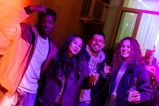 A group of four friends is celebrating together in a vibrant lounge. They are smiling, dancing, and holding drinks in a festive atmosphere filled with colorful lights.