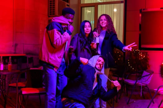 Four friends are gathered outdoors in a lively atmosphere, enjoying drinks and laughter together. The ambiance is filled with colorful lighting and autumn vibes, creating a fun celebration.
