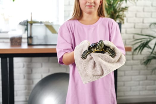 A young child gently cradles a small turtle wrapped in a soft towel. They are indoors, surrounded by plants and a fitness ball, creating a cozy atmosphere.