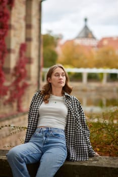 A stylish young woman with wavy brown hair and blue eyes sits on a stone ledge in a historic European city park, wearing a trendy black-and-white plaid shirt, a white crop top, and classic blue jeans. The soft natural lighting highlights the warm autumn atmosphere with golden leaves, red ivy, and medieval architecture in the background. Her relaxed pose and thoughtful expression create a cozy and nostalgic mood, perfect for lifestyle branding, travel promotions, autumn fashion editorials, and advertising seasonal collections. This image captures the essence of autumn in a historic European city, ideal for marketing campaigns, blog posts, and social media content related to travel, fashion, and seasonal themes. Stylish young woman in casual autumn outfit relaxing in historic European city park with medieval architecture, golden leaves, and red ivy, showcasing cozy autumn atmosphere, lifestyle, and travel concepts.