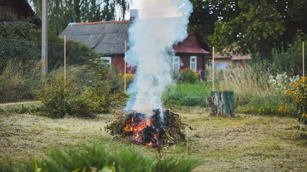 Pile of tree branches burning in a rural garden, producing a large plume of smoke, with a wooden house and fence in the background