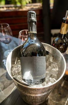 A bottle of champagne stands in an ice bucket, outdoors. The setting suggests a bright celebration, with champagne glasses in the background.
