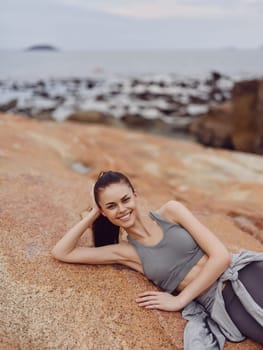 Active woman in gray workout clothing, smiling on a rocky shoreline, promoting fitness, summer energy, and a vibrant lifestyle near water