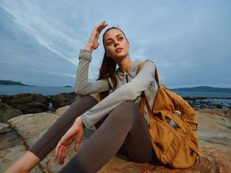 A serene young woman in activewear sits on a rocky shoreline, reflecting on her surroundings, with a backpack beside her, symbolizing adventure and tranquility