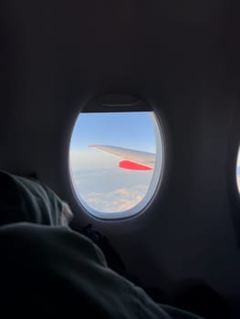 View from airplane window showing wing above clouds during peaceful flight, with passenger resting in seat, soft light creating calm travel atmosphere