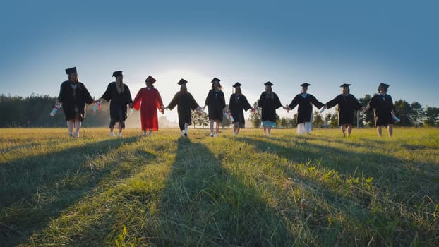 Group of graduates is walking in a field at sunset, holding hands and their diplomas