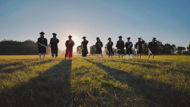 Group of graduates wearing academic gowns and holding diplomas, standing in a field at sunset, celebrating their academic achievement