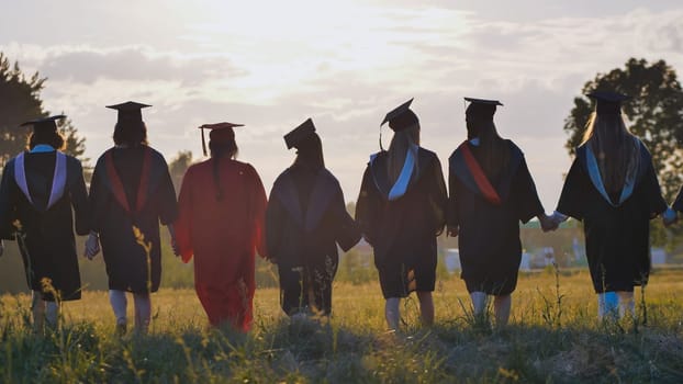 Group of graduates wearing gowns and caps walking together in a field at sunset, holding hands, celebrating their academic achievement and friendship