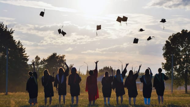 Group of joyful students in academic gowns celebrating graduation by throwing their caps high into the air against a stunning sunset backdrop