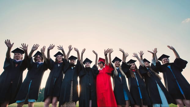 Group of cheerful students wearing graduation gowns and caps waving hands in the air, celebrating their academic achievement outdoors