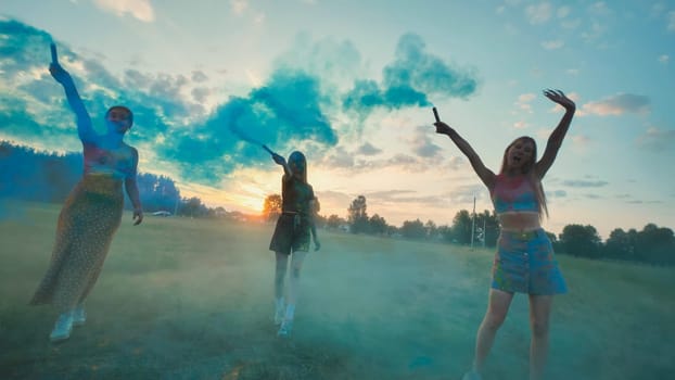 Three young women, probably classmates, joyfully celebrate with blue smoke bombs in a field at sunset, creating a colorful and lively atmosphere