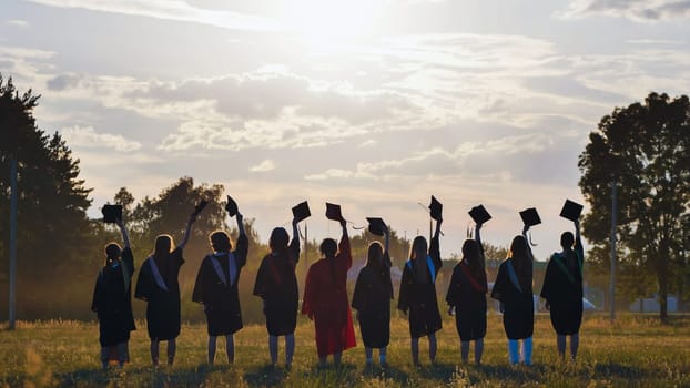 Group of graduates raising their mortarboards in a field at sunset, celebrating their academic achievement and the end of their studies