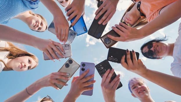 Group of school friends holding smartphones making circle with devices in sky background, low angle view