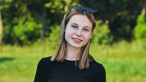 Portrait of a smiling blonde teenage student wearing a black t shirt and sunglasses on her head in a green park