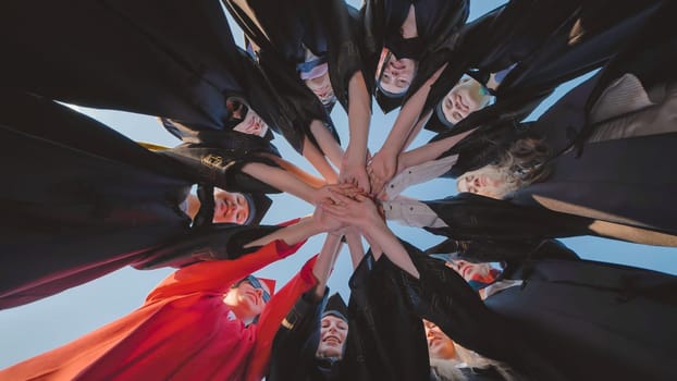 Group of smiling graduates wearing academic gowns joining hands in a circle, celebrating their academic achievement together, viewed from below