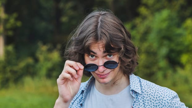 Portrait of a young man in a park, raising his sunglasses and giving a sly smile while making eye contact