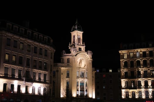 Nighttime view of a historic building in Lyon with a prominent clock tower and sculptural facade, lit by soft architectural lighting.