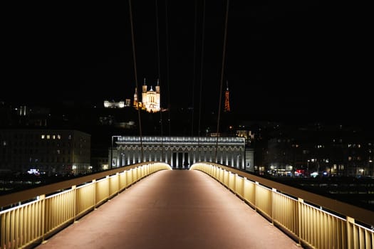 Symmetrical view of a lit pedestrian bridge at night in Lyon, facing the Palais de Justice with the Basilica and tower on the hill.