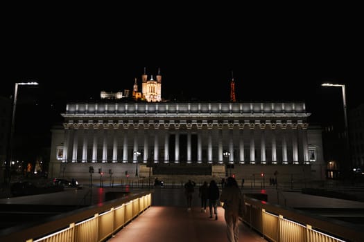 People walking across illuminated bridge at night with Palais de Justice and Basilica of Fourviere glowing on hill in the background. Lyon. France 04.20.2025