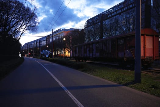 View of a rusted cargo train parked on old tracks beside a quiet street lined with trees and modern buildings during twilight.