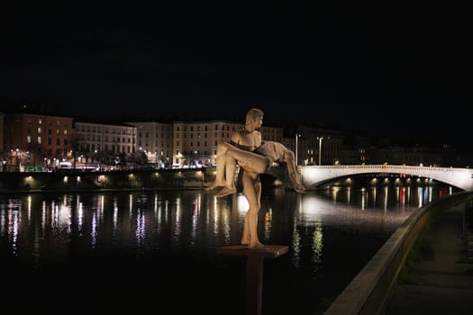 Lyon. France 04.20.2025 Nighttime view of a dramatic statue near a calm river, with city buildings and a lit bridge reflecting in the water.