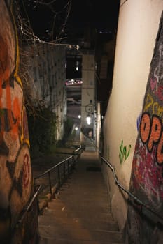 Lyon. France 04.20.2025 Night view of a narrow, graffiti-covered stairway descending through an urban alley, illuminated by soft street lamps and city lights.