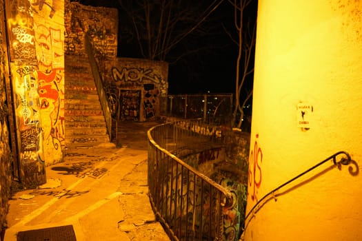 Nighttime view of a vibrant, graffiti-filled staircase and walkway in an urban setting, lit by warm artificial lighting.
