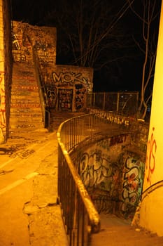 Nighttime view of an outdoor urban staircase spiraling downward, fully covered in colorful graffiti and dimly lit by orange streetlight.