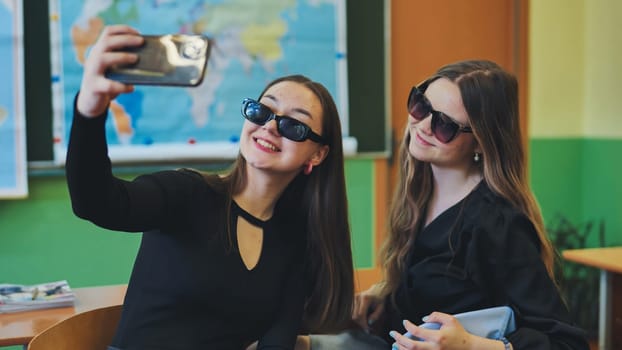 Two cheerful female students wearing sunglasses are taking a selfie with a smartphone in a classroom