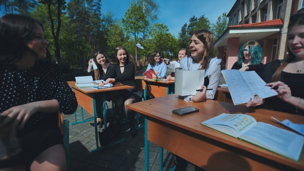 Group of cheerful teenagers enjoying their school day, studying and chatting together at desks outside on a sunny day