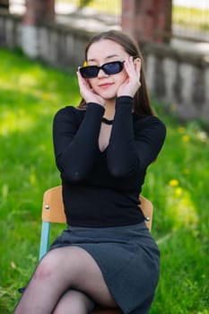 Female student taking a relaxing break outdoors, sitting on a chair in a sunny park, sporting sunglasses on a beautiful school day