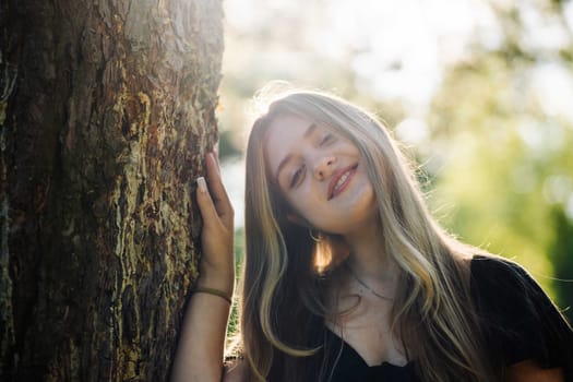Portrait of a smiling blonde teen girl leaning against a tree trunk in a park during a sunny summer day