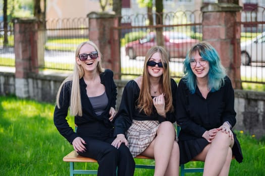 Three cheerful students wearing sunglasses are laughing and enjoying their time together while sitting on a bench in the schoolyard during a break