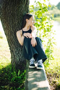 Serene female relaxing near lakeside tree, finding calm amid lush summer landscape, connecting with natural surroundings