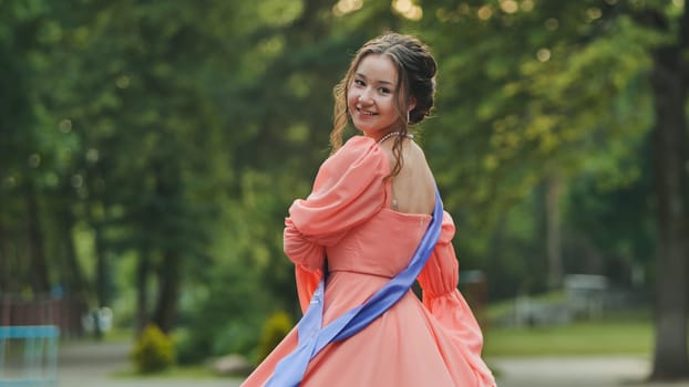 Graduate student smiling brightly while wearing a sash, joyfully turning around in a lush park, celebrating graduation and future achievements