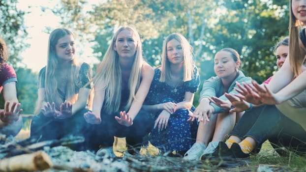 Group of young female students warming their hands by the bonfire during their summer camp vacation in the forest