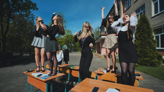 Five cheerful high school girls dancing on school desks in the schoolyard, celebrating the end of the school day