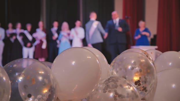 Filling the foreground, white and transparent balloons with gold and silver confetti contrast with blurred figures of graduating students and teachers on stage