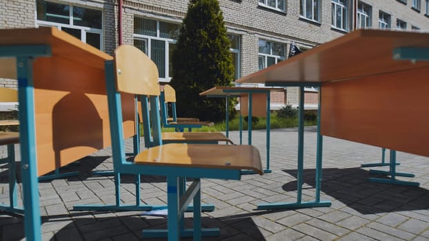 Empty school desks and chairs stand outdoors on a paved area in front of a school building, suggesting a break or unusual class setting during the school day