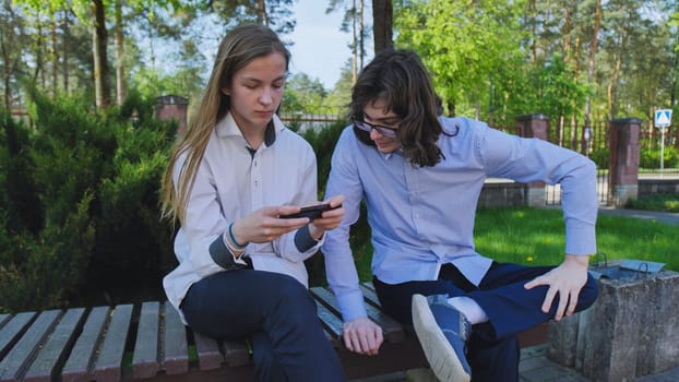 Two high school students are sitting on a bench outdoors, playing a mobile game together during their school break