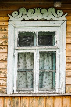 Weathered window with peeling paint, carved valance, and floral curtains, adding a touch of nostalgia to a traditional wooden house