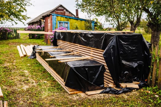 Stacks of wooden planks covered with black plastic sheet material protecting from rain near colorful rural house in countryside